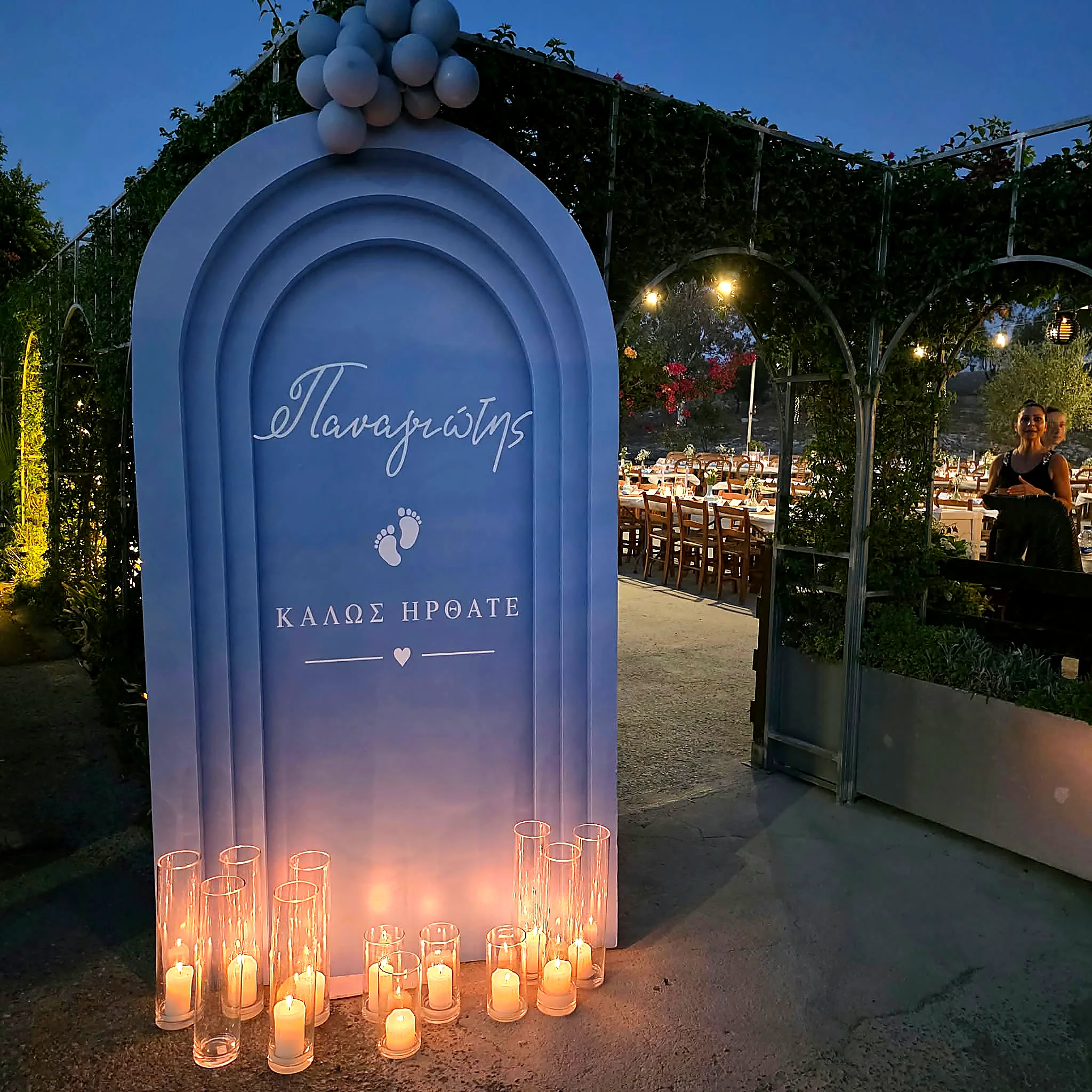 A pale blue christening entrance arch lit by candles in a garden at night, with a welcome message