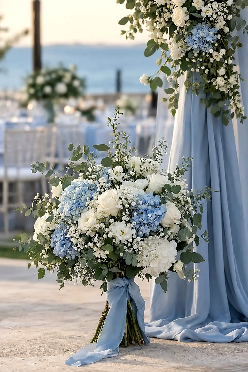 A bridal bouquet of white roses and blue hydrangeas beside a draped column overlooking the sea