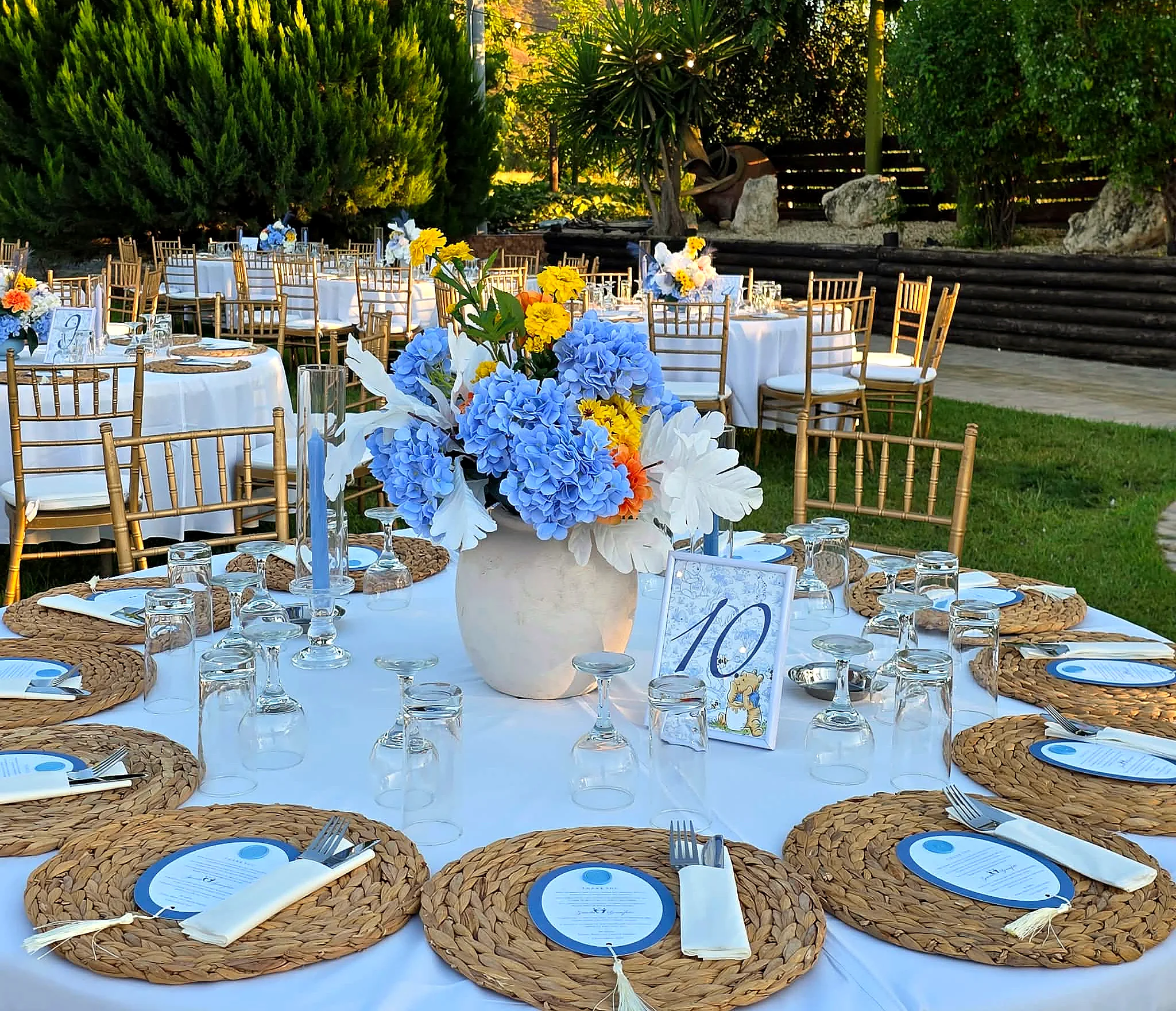 A garden table set with blue and yellow florals, woven placemats and gold chiavari chairs