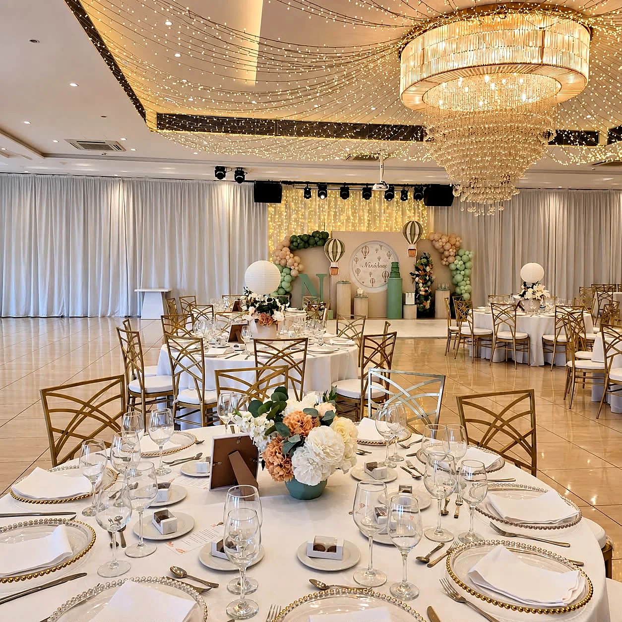 A ballroom reception table laid in white beneath a crystal chandelier and a fairy-light ceiling