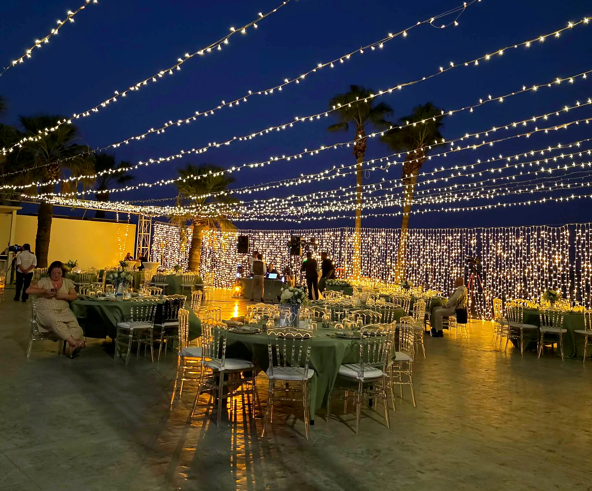 An evening reception beneath canopies of festoon lights with palm trees against a deep blue sky