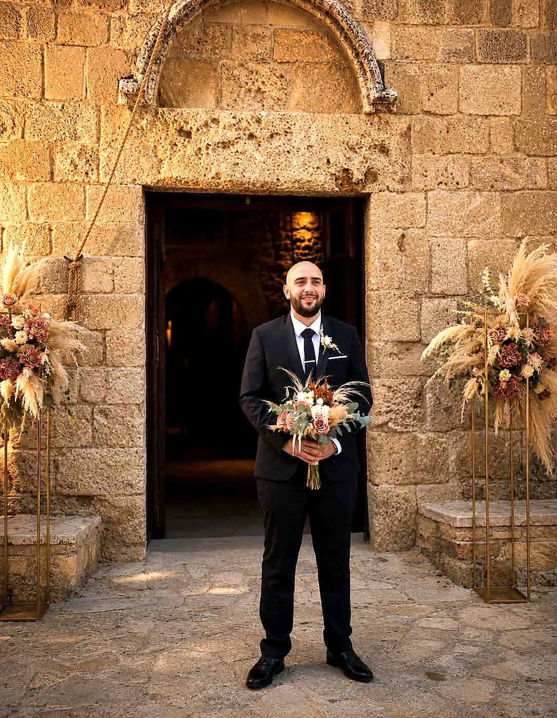 A groom standing in a stone church doorway framed with white florals