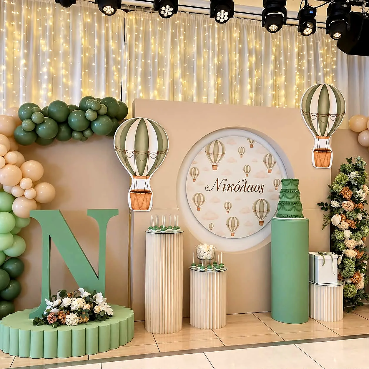 Sage green plinths, balloon garlands and hot-air-balloon props against a fairy-light curtain