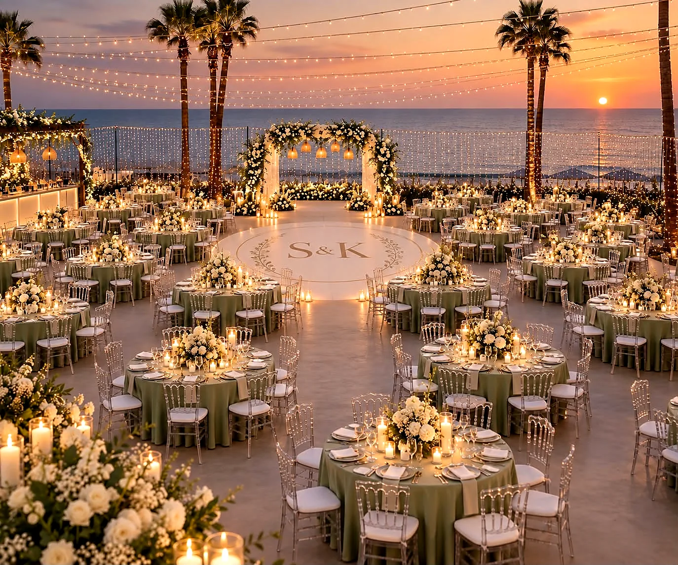A seaside reception at dusk with lit palms, a floral ceremony arch and tables set in white